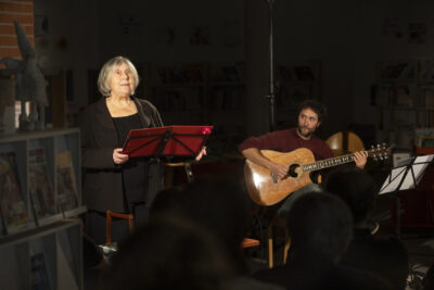 dans la médiathèque plongée dans la semi-pénombre et la lumière tamisée, une comédienne aux cheveux blonds vêtue d'un costume noir se tient debout derrière un pupitre rouge pourpre tandis qu'un guitariste assis apparaît sous le spot de lumière en arrière-plan - Agrandir l'image 1 sur 3, fenêtre modale