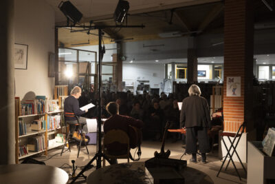 Dos aux musicien et comédiens sur scène, la médiathèque avec ses rayonnages apparaît dans une ambiance nocturne éclairée de spots tandis que le public se devine en arrière-plan dans la pénombre - Agrandir l'image 2 sur 3, fenêtre modale