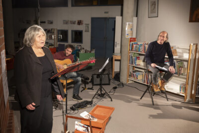 photo d'ensemble des artistes de la compagnie Entresort sur une scène improvisée entre les rayonnages de la Médiathèque, à savoir deux comédiens, une femme debout en train de déclamer à gauche, un homme juché sur un tabouret de bar à droite, chacun devant leur un pupitre de part et d'autre de l'image, accompagné à l'arrière-plan d'un guitariste en train d'égrener ses notes dans un geste délicat - Agrandir l'image 3 sur 3, fenêtre modale