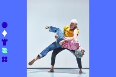Image de danse en duo un danseur debout tenant un autre danseur plus jeune à bout de bras tandis que ce dernier rejette son corpsen arrière en confiance. la scène est lumineuse, les vêtements jeans et tee-shirt, frais et colorés