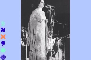 photo en noir et blanc des années 60 d'une chanteuse orientale de renom (Warda) sur une scène d'orchestre debout au micro en robe claire surmontée de broderies