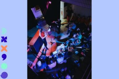 photo en plongée du parterre de public rassemblé autour d'une prestation musicale, de flûtiste debout au micro dans une salle plongée dans l'obscurité et les éclairages bleu et rose