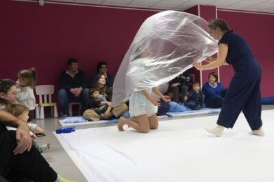 Devant le public familial assis tout autour, sur un coin de la scène drapée de blanc du sol de la maison de quartier de la Paderne, une jeune danseuse de short bleu pâle vêtue, s'agenouille sous la voile transparente qu'agite l'autre comédienne debout à droite de l'image l'enveloppant dans cette bulle qui figure la toute petite vague dont le spectacle est le thème - Agrandir l'image 3 sur 5, fenêtre modale