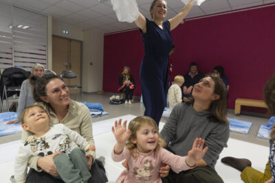 Une fmille accroupie sur la sc-ène mamans et fillettes s'émerveillent du décor de théâtre qui virevolte au-dessus d'elles entre les mains d'une danseuse de la compagnie Paracosm debout au-dessus d'elle - Agrandir l'image 5 sur 5, fenêtre modale