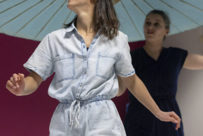 debout d'une démarche gracieuse une "mère" vêtue d'une combinaison légère de couleur sombre qui la fait passer pour une silhouette bienveillante en arrière-plan, et sa "fillette" incarnée par une danseuse d'âge adulte, la mine émerveillée, s'avancent sous une ombrelle chinoise en papier de riz bleu pâle et arceaux en bois léger face caméra à l'intérieur de la salle de la maison de quartier de la Paderne. - Agrandir l'image 2 sur 5, fenêtre modale