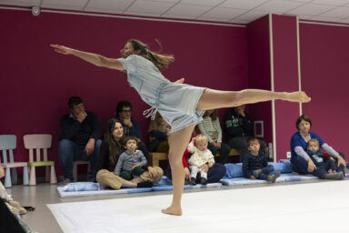 une danseuse incarnant une fillette vêtue d'un baigneur bleu pale s'lénce dans un mouvement très enfantin et très dynamique de danse main gauche tendue vers la gauche de l'image sur une scène couleur crème face au public réuni à la maison de quartier de la paderne