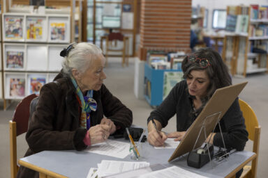 Assise à une table de la médiathèque une dame distinguée aux cheveux blancs ramassés en chignon, manteau en daim marron et foulard coloré dans les cheveux, écoute une médiatrice attablée à ses côtés, chevux longs bruns lâchés et regard de cendre baissés sur son travail d'écriture au stylo bic.
