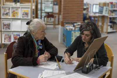 Assise à une table de la médiathèque une dame distinguée aux cheveux blancs ramassés en chignon, manteau en daim marron et foulard coloré dans les cheveux, écoute une médiatrice attablée à ses côtés, chevux longs bruns lâchés et regard de cendre baissés sur son travail d'écriture au stylo bic. - Agrandir l'image 1 sur 14, fenêtre modale