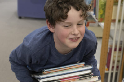 Entre deux rayonnages, un jeune garçon souriant en train de porter une pile de livres jeunesse entre ses bras, fait mine d'être accablé sous le poids sous le regard du photographe - Agrandir l'image 8 sur 14, fenêtre modale