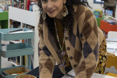Une jeune fille pétillante maquillée dans un style oriental, pendentifs dorés ouvragés aux oreilles, se penche sur un plan d etravail où elle est en trainb d'apposer un collage coloré de pigeon sur une photo en noir et blanc et sourit au photographe. - Agrandir l'image 2 sur 14, fenêtre modale