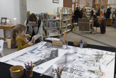 Une fillette blonde à queue de cheval se fait aider de sa mère dans la réalisation d'un collage coloré qu'elle applique sur une planche photographique en noir et blanc de vues de la Médiathèque - Agrandir l'image 3 sur 14, fenêtre modale