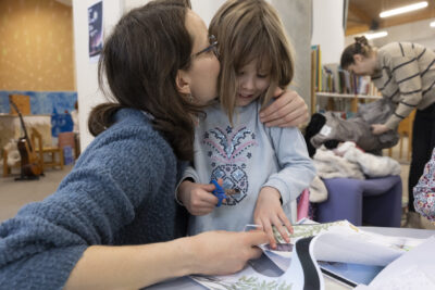 scène de tendresse et de complicité entre une mère et sa fille, toutes deux vêtues de bleu, en train de participer à un atelier créatif leurs bras et mains de la même carnation rosée. A l'arrière-plan la vie d'une médiathèque, guitare apposée contre un mur en chantier, femme en train de déposer son manteau sur une chauffeuse. - Agrandir l'image 5 sur 14, fenêtre modale