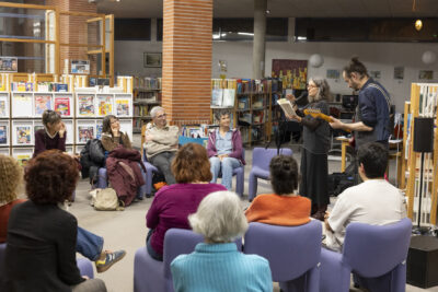 A droite, deux comédiens guitariste et lectrice , s'adressent au public installé dans des chauffeuses couleur parme au milieu des rayonnages de la médiathèque. Les couleurs des magazines, des vêtements et des tableaux accrochés aux murs, rehaussent la vue d'ensemble. - Agrandir l'image 13 sur 14, fenêtre modale