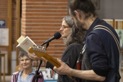 Plan rapproché sur une lectrice professionnelle de profil, lunettes à montures rouges sur le nez, en train de lire au micro un extrait de Les Pays de Marie-Hélène Lafon, accompagnée au premier plan d'un guitariste en bras de chemises, cheveux longs attachés en catogan; en arrière-plan flouté, le visage au sourire complice d'une dame du public. - Agrandir l'image 14 sur 14, fenêtre modale