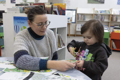 au centre de rayonnages, une mère en pull de laine avec plusieurs tons de bleu et beige, montures à écaille, assiste sa très jeune fille aux cheveux fins, l'air appliqué dans sa tâche de découpe d'une image colorée, un sweat à l'effigie des bisounours, personnages de dessin animé, aux abords d'une table de travail créatif. - Agrandir l'image 6 sur 14, fenêtre modale