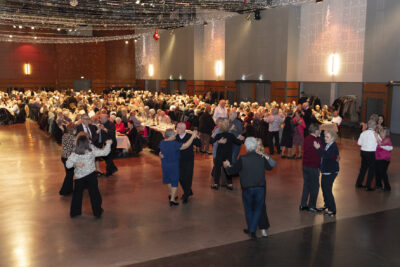 Photo de la salle du Phare dans laquelle de grandes tables sont disposées avec des les séniors en train de manger et une quinzaine au premier plan qui dansent en slow - Agrandir l'image 20 sur 20, fenêtre modale