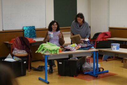 photo de deux femmes dans une salle, devant une malle avec plein d'objets répartis dedans - Agrandir l'image, fenêtre modale