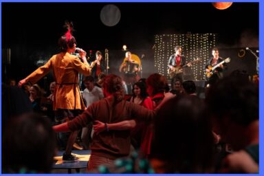photo de nuit mêlant scène et public aux tenues chamarrées réunis dans l'ambiance festive d'un concert éclairé par les lumière de scène, les musiciesn en costumes debout au micro sont disposés en rond autour du public en train de danser dans une chorégraphie collective, bras écartés à hauteur des hanches, de dos à l'objectif.