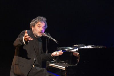 assis devant son piano Yamaha joliment souligné de rais de lumière bleu néon, cheveux en broussaille et barbe de trois jours, Babx pivote à 90° en direction du public pour s'adresser à lui en parlant avec les mains d'un air interrogateur. - Agrandir l'image 12 sur 13, fenêtre modale