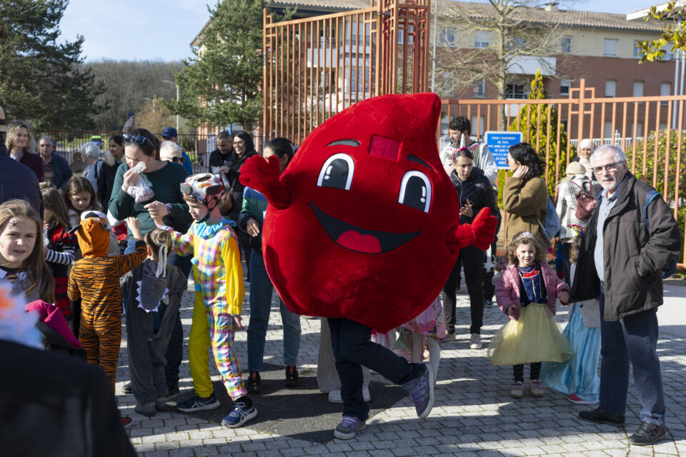 photo de familles sur la place en extérieur, au soleil, avec la mascotte du don du sang qui prend la pose pour l'objectif