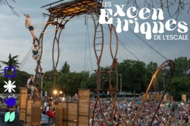 vue d'ensemble depuis larrirère-scène d'un podium installé dans un espace vert à la tombée de la nuit, le public assis dans le parc rassemblé autour de la scène surmontée d'une plateforme acrobatique en bois, les circassiens en justaucorps se balançant en hauteur sur leurs agrès
