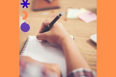 Plan rapproché sur les avant-bras d'une jeune fille en manches de chemise à carreaux entrain d'écrire au stylo plume dans un cahier à spirale sur un plan de travail en bois naturel jonché de feuilles de papier, l'image est floûtée et lumineuse en tons de beige et rosé