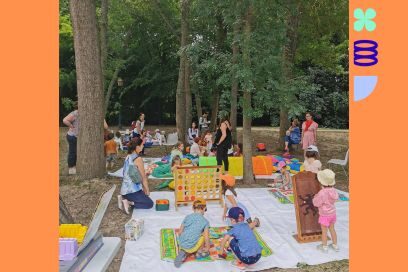 Modules en bois colorés, livres jeunesse, jeux, éparpillés sur une gigantesque nappe de pique-nique dans un parc entouré d'arbres, adultes debout et enfants installés au sol sont affairés dans une joyeuse animation. - Agrandir l'image, fenêtre modale