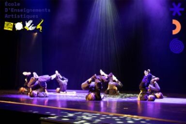 photo prise sur scène pendant une séquence de spectacle de danse hip hop, les danseurs au sol dans une position acrobatique, en appui sur la tête dans une lumière violette, leurs postures éclairées d'en haut par les projecteurs tamisés