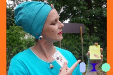 Vêtue de bleu cyan, un foulard noué sur la tête, le profil grave et des amulettes ésotériques autour du cou et des oreilles, une dame indique due la main droite une carte à jouer qui représente un légume dans sa main gauche, dans un décor extérieur de verdure