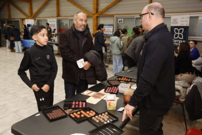 photo d'un jeune garçon et d'un homme qui échangent avec un homme debout derrière un stand sur lequel sont posées des boîtes de chocolats - Agrandir l'image 2 sur 22, fenêtre modale
