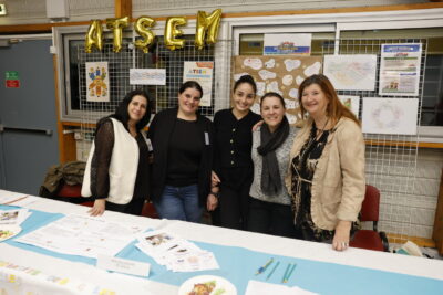 photo de cinq femmes debout derrière un stand qui posent et sourient à l'objectif, au dessus derrière elle, le signe "ATSEM" - Agrandir l'image 9 sur 22, fenêtre modale