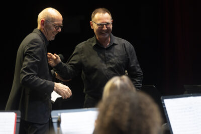 Plan rapproché sur les deux directeurs musicaux souriants, respectivement de l'Orchestre d'Harmonie et des Sourds-Doués, debout en costume et chemise noirs et lunettes à montures dorées. - Agrandir l'image 7 sur 19, fenêtre modale