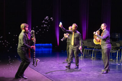 Le quatuor sur la scène nimbée de lumière violette, la saxo à gauche s'avançant face aux trois autres, dont celui du milieu diffuse des b=ulles de savpon dans l'air autour d'eux composant une image esthétique. - Agrandir l'image 2 sur 19, fenêtre modale