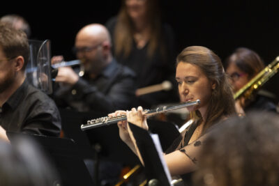 Plan rapproché sur une flûtiste aux cheveux châtains longs et attachés en robe de voile noir brodée, en train de jouer de son instrument au milieu du parterre des musiciens floûté. - Agrandir l'image 8 sur 19, fenêtre modale