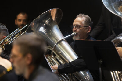 Plan rapproché sur le joueur de tuba, au métal mat et argenté, le musicien en train de souffler dans son instrument, arbore une une barbe grisonnante et des lunettes cerclées de métal, on devine qu'il se trouve au milieu des autres musiciens. - Agrandir l'image 11 sur 19, fenêtre modale