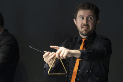 Plan rapproché sur l'un des musiciens des Sourds-Doués en chemise noire et cravate orange fine, en train de jouer d'un triangle doré, il est châtain-blond aux yeux bleus et petite barbe soignée, les yeux tournés hors champ vers la droite de l'image. - Agrandir l'image 12 sur 19, fenêtre modale