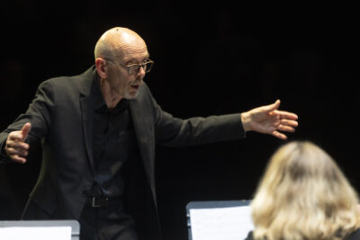 Debout, bras ouverts, crâne rasé et nez chaussé de lunettes, le chef d'orchestre Claude Puysségur apparaît sur la ligne claire de la scène en pleine concentration, la mine expressive, face à ses musiciens que l'on devine au premier plan. - Agrandir l'image 14 sur 19, fenêtre modale