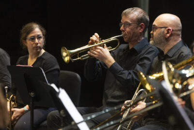 Pendent le concert, plan rapproché sur le joueur de trombone doré en train de jouer un solo assis derrière son pupitre, cheveux courts grisonnants et petites lunettes de métal rondes, , entouré d'autres musiciens à vent tournés vers lui. - Agrandir l'image 15 sur 19, fenêtre modale