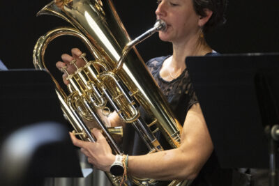 Plan rapproché sur une joueuse de tuba doré, tenant fermement son instrument de ses bras musclés, cheveux courts elle apparaît les yeux baissés sur sa partition, en petite robe noire assise de manière souple derrière son pupitre. - Agrandir l'image 16 sur 19, fenêtre modale