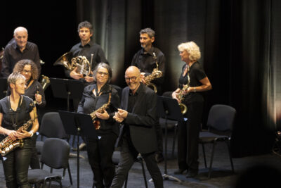 Tout sourire, sur scène encadrée de rideaux gris en velours, le directeur musical de la Société Musicale de Tournefeuille, svelte, crâne rasé, en costume cintré anthracite, s'avance au milieu de ses musiciens debout en se frottant les mains. - Agrandir l'image 3 sur 19, fenêtre modale