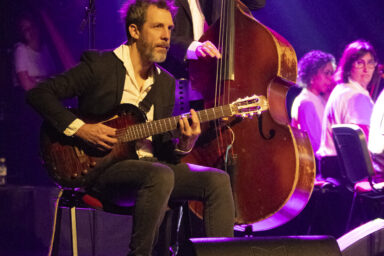 Plan rapproché sur deux musiciencs de Samarabalouf debout, contrebasse et guitare classique, vêtus de chemise blanche à col pointu et veste de blazer noire, les yeux souriants, au bord de la scène dans la lumière violette.