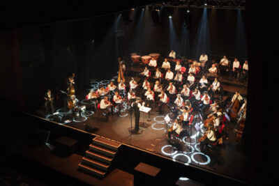 Vue d'ensemble de l'estrade pendant le concert avec ses marches d'accès depuis le public et sur l'orchestre dans la lumière cuivrée, des cercles blancs de lumière jonchant la scène habillant la scène. - Agrandir l'image 7 sur 19, fenêtre modale