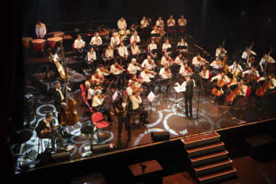 Vue partielle de la scène avec à gauche le trio de jazz manouche Samarabalouf accompagnant l'orchestre symphonique à droite sous la direction de Claude Puysségur debout de dos au public en manches de chemise blanches. - Agrandir l'image 8 sur 19, fenêtre modale