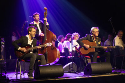 Le trio Samarabalouf, la contrebasse debout, les guitaristes sylvain Peyrières et François Petit assis sur le devant de scène, mines expressives en arrière-plan les musiciens de l'orchestre symphonique apparaissent en chemise blanche dont certains orientent le regard vers le trio dans la lumière bleue. - Agrandir l'image 2 sur 19, fenêtre modale