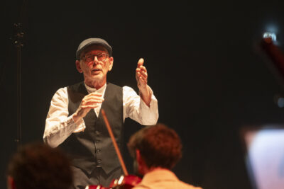 Plan rapproché sur le directeur musical Claude Puysségur, fendant l'air devant son visage concentré en veston et casquette de Gavroche sur une chemise blanche. Ses traits sont rehaussés d'ombres qui le font apparaître comme croqué sur un dessin. - Agrandir l'image 9 sur 19, fenêtre modale