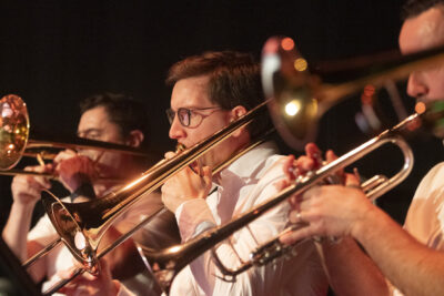 Gros plan sur les trombonistes en plein jeu dans la lumière alignés en diagonale de gauche à droite de l'image dans un mouvement parfaitement coordonné - Agrandir l'image 11 sur 19, fenêtre modale