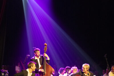 Regard complice entre Sylvain Peyrières et François Petit, guitaristes du trio Samarabalouf, jouant avec le contrebassiste derrière eux, sur l'avant-scène les musiciens de l'orchestre symphonique apparaissant en arrière-plan sous les rayons violets des projecteurs. - Agrandir l'image 3 sur 19, fenêtre modale