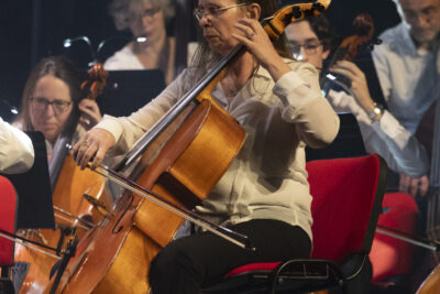 Gros plan en teintes crème rouge et noir, sur la violoncelliste lunettes rondes et cheveux châtains attachés, pantalon noir et chemise blanche, au milieu des autres musiciens tous en pleine action, avec effet de mouvement qui floûte l'image. - Agrandir l'image 13 sur 19, fenêtre modale