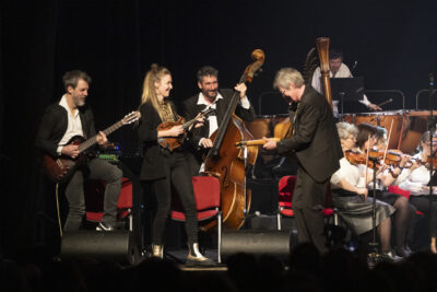 scène de complicité au sein de la formation de Samarabalouf pendant le concert, tous les musiciens, deux guitaristes, une mandoline et la contrebasse debout jouant en se regardant les uns les autres tout sourire, en costume noir. - Agrandir l'image 15 sur 19, fenêtre modale