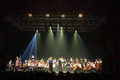 Vue d'ensemble des musiciens sous les lumières fumées et irisées de vert-bleu, la chanteuse Corinne Champy en chemisier bleu canard se tient debout à côté du chef d'orchestre Claude Puyssegur de dos en train de diriger ses musiciens, à gauche le quatuor Samarabalouf. - Agrandir l'image 16 sur 19, fenêtre modale